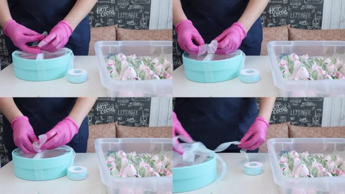 一位妇女正在为自制的棉花糖做包装。她试了试青绿色盒子的丝带长度。她用剪刀剪丝带。桌子上有西风郁金香。