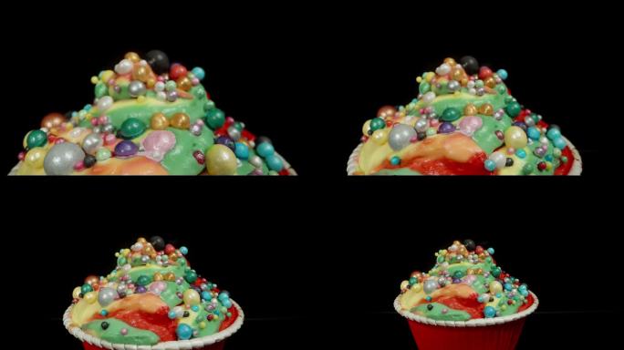 一个杯子蛋糕，有五彩缤纷的彩虹糖霜和球，在黑色的背景上孤立。多莉滑块，近一点。