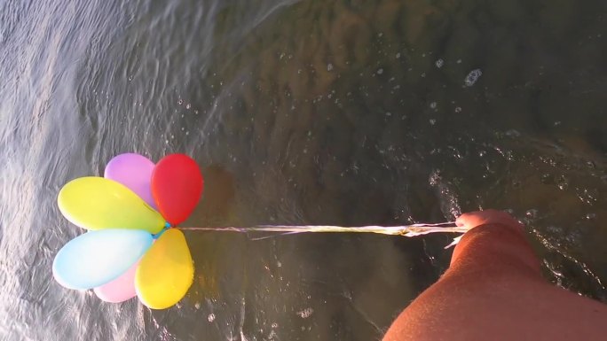 在一个阳光明媚的早晨，一个人拉着一束彩色的小气球，用丝带绑在水面上。慢动作