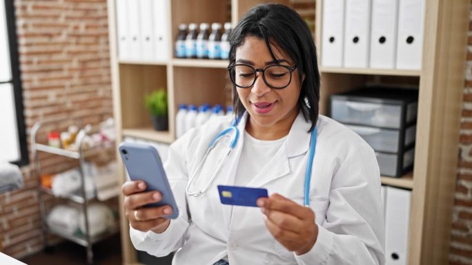 一位微笑的西班牙裔女性，身穿白大褂，在诊所内部使用智能手机和信用卡