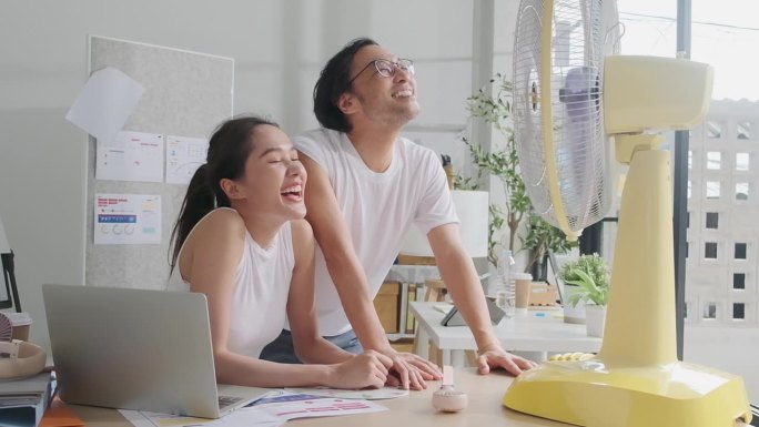 夏天，在一间小办公室里，两名亚洲员工正在用风扇吹风。