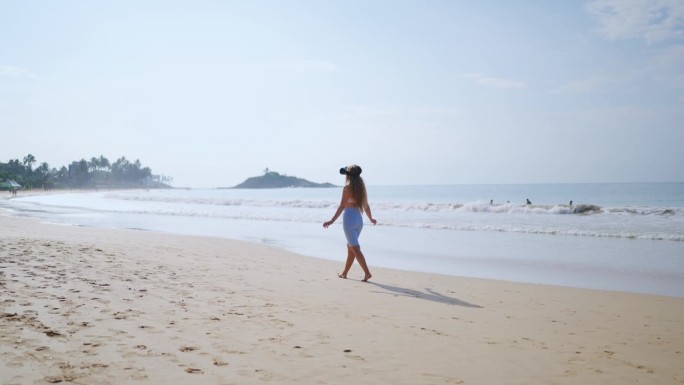 女人在海滩上通过VR头盔体验虚拟热带天堂。沿着海岸散步，波浪轻轻地拍打着附近。交互式技术将用户带入数