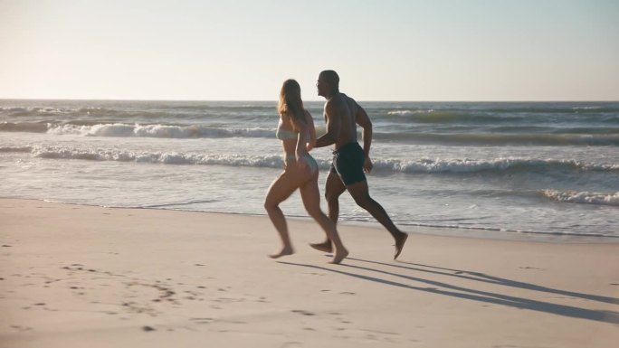 一对穿着泳装度假的情侣手牵着手沿着海滩岸线奔跑
