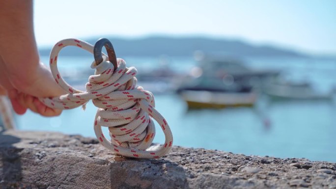 一个人用一根红白相间的卷绳固定一个锚，背景是风景优美的海景和停泊的船只