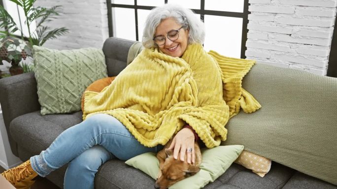 一位白发苍苍、披着黄毯子的妇女和她的宠物狗坐在室内的沙发上微笑着