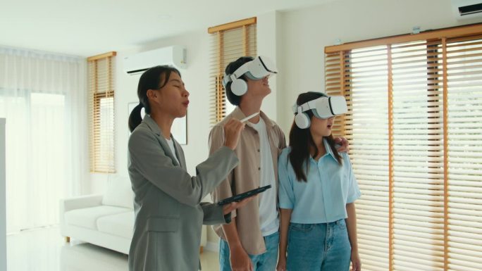 亚洲女房地产经纪人在新房子里用VR和平板电脑向租客解释和展示租或买。房东把房产卖给幸福夫妇客户。
