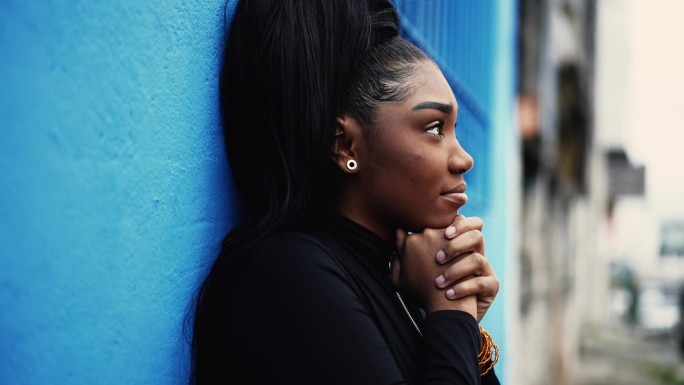 一位满怀希望的年轻黑人妇女站在城市街道外，带着信念和感激向上凝视。非裔美国成年女孩在城市环境中抬头微