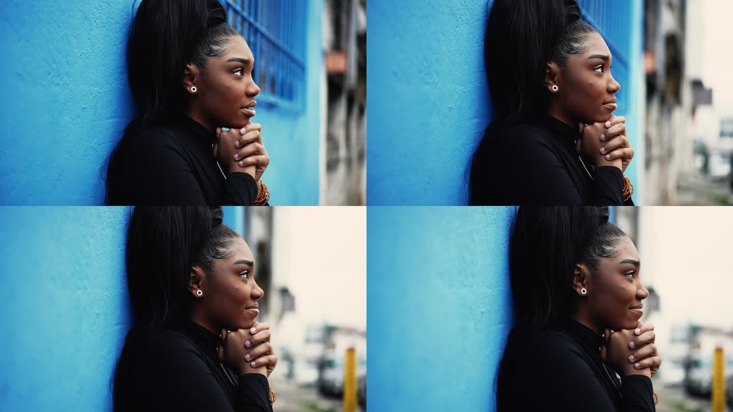 一位满怀希望的年轻黑人妇女站在城市街道外，带着信念和感激向上凝视。非裔美国成年女孩在城市环境中抬头微