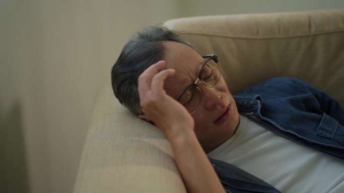 亚洲老人感到疲惫，躺在客厅的沙发上，白发老人痛苦，压力，年龄问题，伤害和孤独，退休后的心理健康问题，