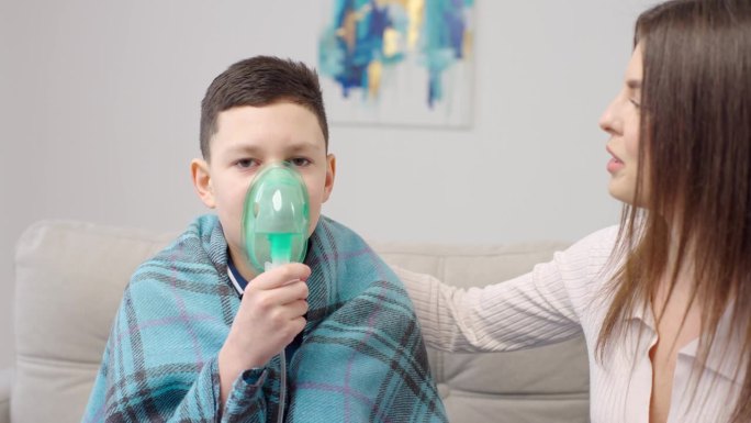 母亲在家里的沙发上拥抱生病的儿子，帮他使用喷雾器。女人用器械给男孩吸气