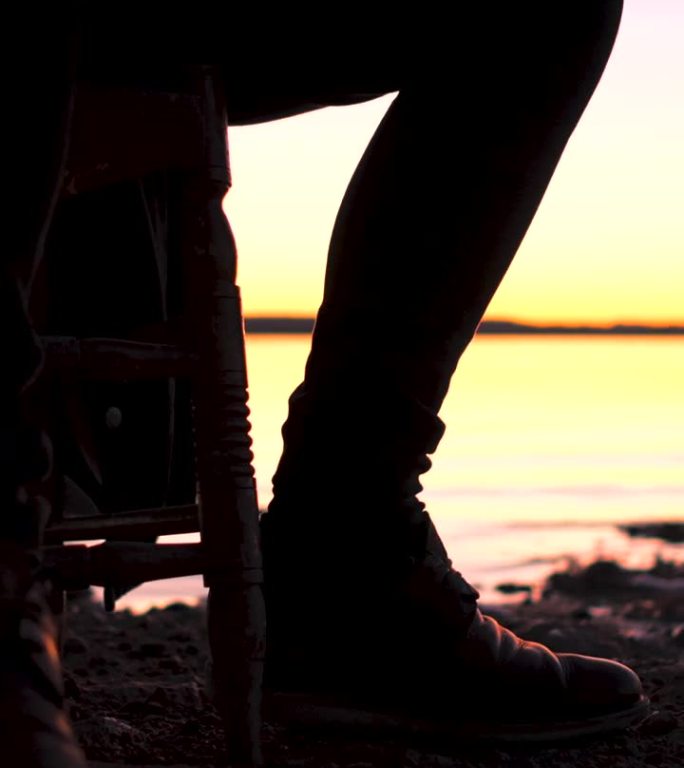 日落时分，潮人在湖边弹吉他的剪影。一名吉他手坐在椅子上，用腿敲打着拍子，在黄昏时分用原声吉他即兴演奏