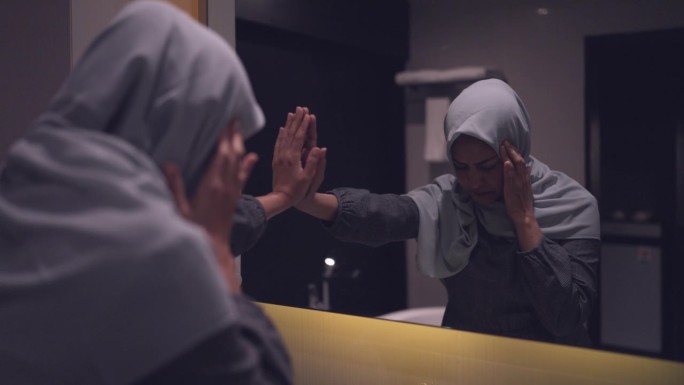 一位戴着头巾的妇女站在浴室的镜子前，对家庭生活感到压力很大。