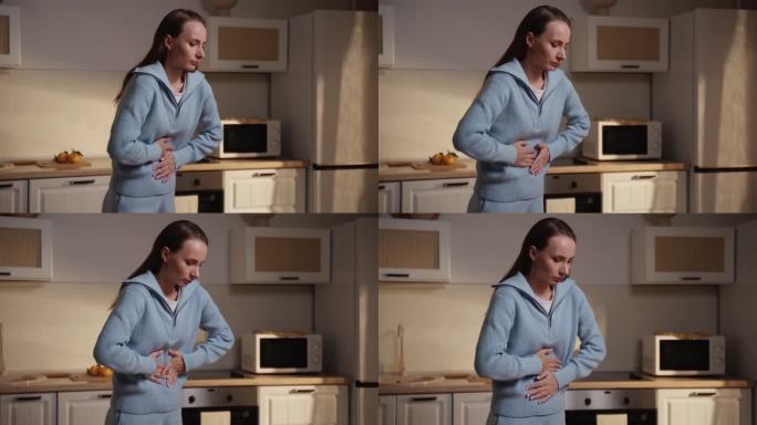 年轻女子在家里胃痛。一个穿蓝色毛衣的女人站在厨房里，痛苦地捂着肚子