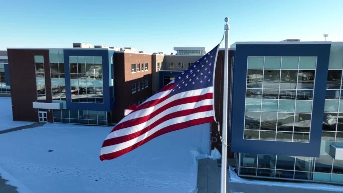 美国国旗的空中特写。学校建筑的背景与明亮的早晨的阳光。地上有雪。