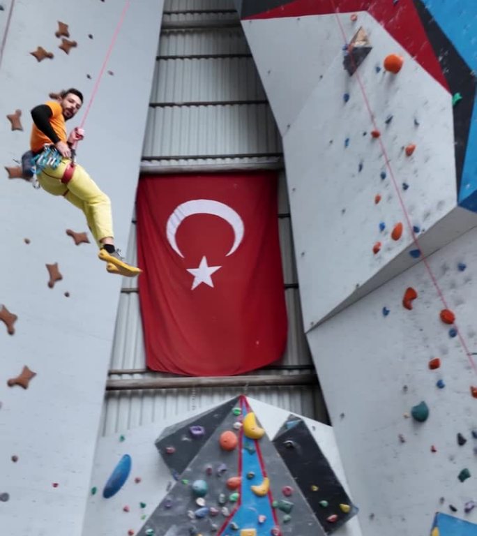 男攀岩者用系在抱石墙上的攀岩绳荡来荡去。墙上挂着彩色的巨石握把。