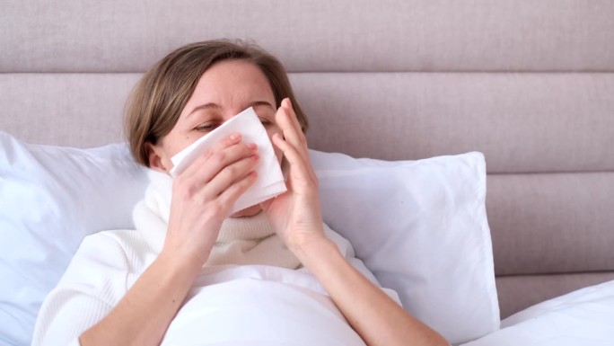 一名年轻女子在卧室的白色床上对着纸巾打喷嚏，流感和过敏的概念。