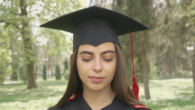 漂亮的黑发女孩刚从大学毕业，站在公园里，眼睛眨着，环顾四周。