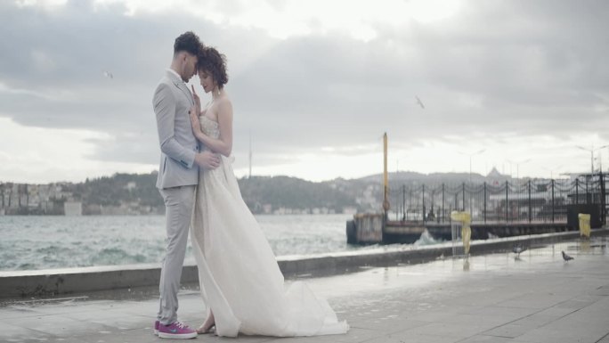 浪漫的新婚夫妇互相拥抱，站在码头上，面对着模糊的城市景观和绿色的山丘。行动。汹涌的海浪和恩爱的新郎新