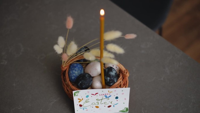 特写镜头。在室内的桌子上，一只燃烧的蜡烛插在一个装饰过的篮子里，篮子里放着复活节彩蛋。篮子旁边有一张