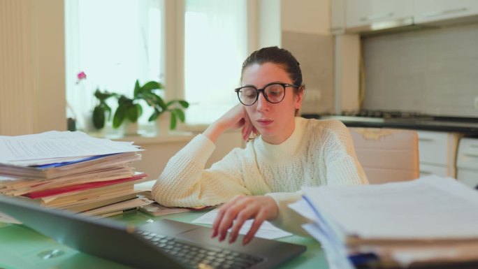 戴眼镜的女人在家里用笔记本电脑工作，桌子上散落着文件