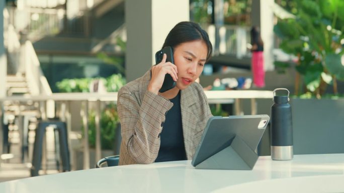 年轻的亚洲女商人一边用平板电脑工作一边打电话，她坐在户外，展示了高效的多任务处理。户外可持续经营。