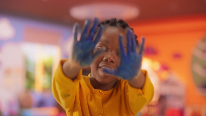 才华横溢的非裔美国女孩在幼儿园的艺术治疗期间用她的手画水彩画。幼儿看镜头，用视觉艺术表达情感