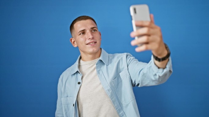 一个微笑的年轻西班牙男子拿着他的智能手机对着室外一堵孤立的蓝色墙自拍。