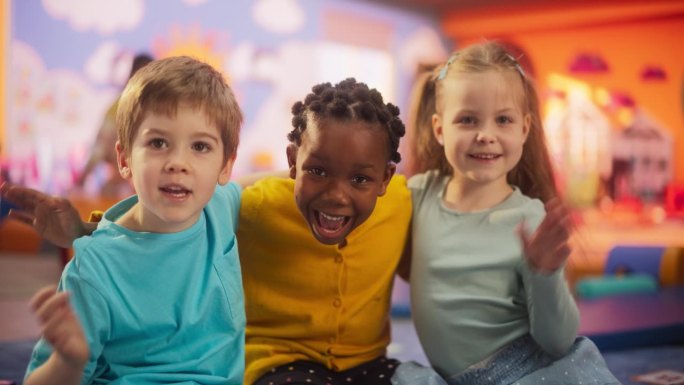 在现代幼儿园里，快乐多样的孩子们坐在地板上一起摆姿势的肖像。美丽的国际小朋友看镜头，微笑，挥手