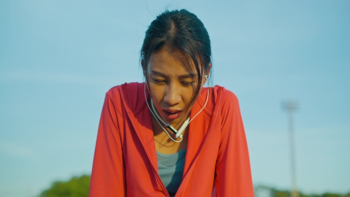 年轻的亚洲女运动员在户外跑步后，疲惫地戴着耳机听着音乐。健康的锻炼。