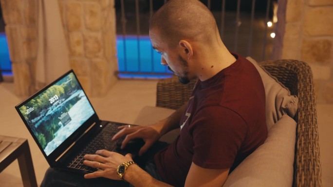 秃顶的年轻自由职业者晚上坐在别墅阳台的沙发上用笔记本电脑工作