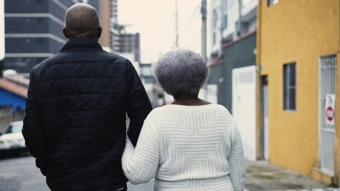 非裔美国成年儿子的背影与白发苍苍的母亲在城市散步，80多岁的老太太与看门人手挽手在户外细雨中