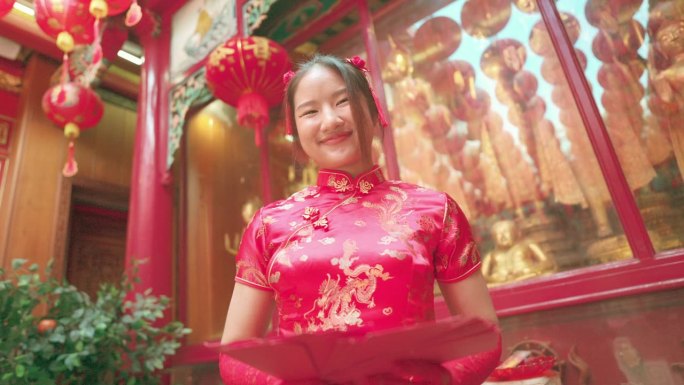 年轻的亚洲女孩穿着红色旗袍，在中国寺庙里拿着红包，向空中扔红包。给父母礼物或钱。