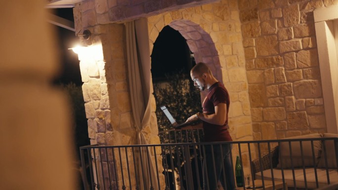 晚上，秃顶的年轻人站在别墅阳台上使用笔记本电脑