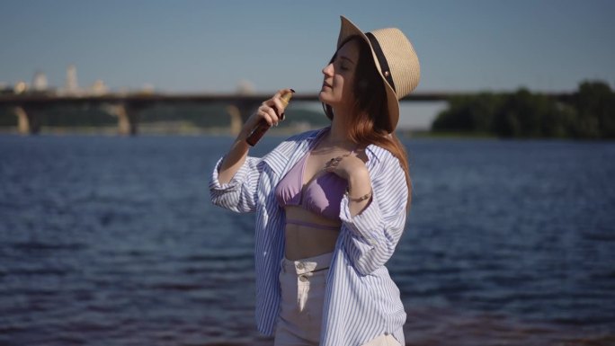 一个穿着衬衫、牛仔短裤、戴着边帽的女孩站在河边的沙滩上跳舞，手里拿着防晒喷雾。