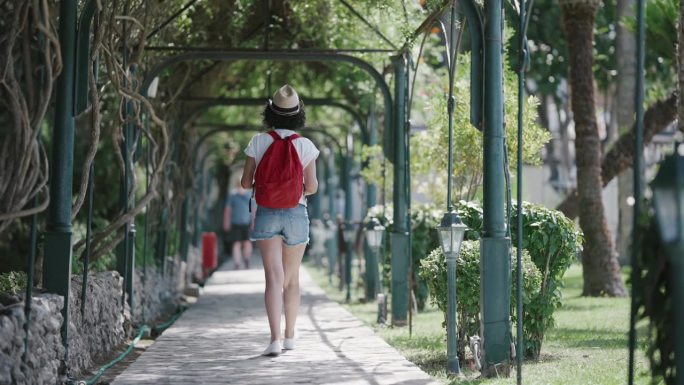 身材匀称的女游客背着背包，头戴草帽，慢镜头漫步在希腊科孚岛上的阿基里翁宫花园