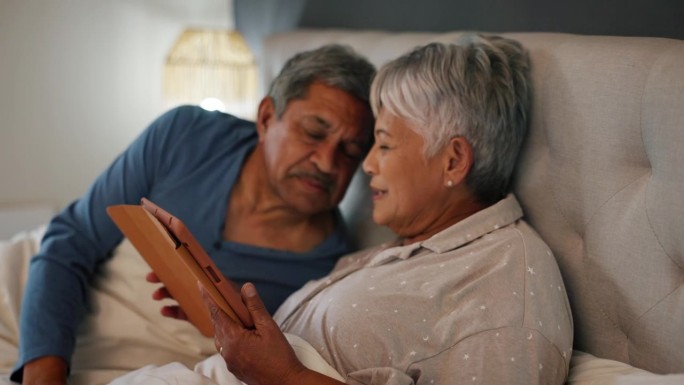 亲吻，平板电脑和长辈在床上玩社交媒体或手机应用，一起放松。爱情，数字技术和老年男女在家里的卧室里的纽