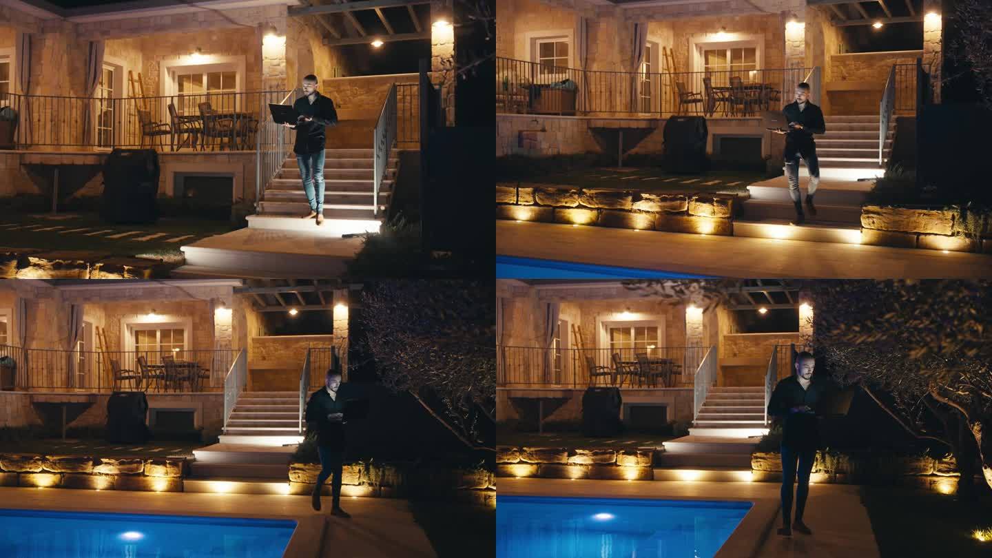 晚上，在有灯光的别墅外的游泳池旁使用笔记本电脑的年轻人