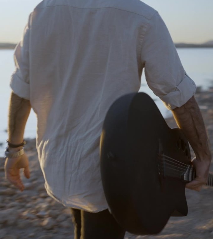 潮人带着吉他欣赏日落在湖岸的石头沙滩上散步对着太阳-慢动作的后景