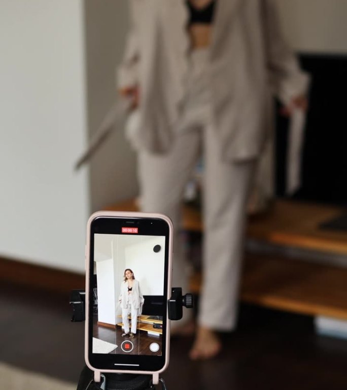 这款手机立在一个三脚架上，拍摄一个博主女孩进入画面并摆姿势的视频。博主在房间里用手机拍摄自己，房间里