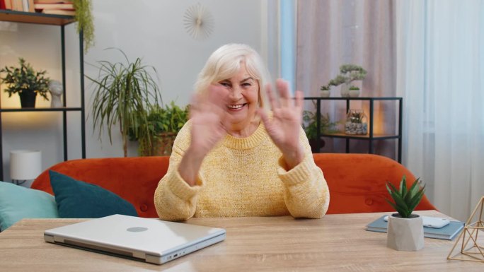 一位年长的女士对着镜头友好地微笑，在家里的沙发上挥手打招呼