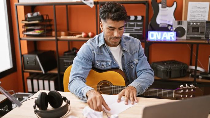 一名西班牙裔男子在音乐工作室沮丧地揉纸，拿着吉他和“正在播放”的牌子。