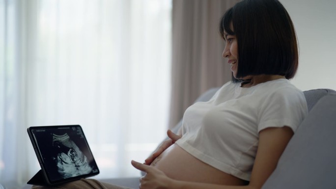 年轻的怀孕母亲高兴地看着平板上的超声波片，与她的孩子交谈。