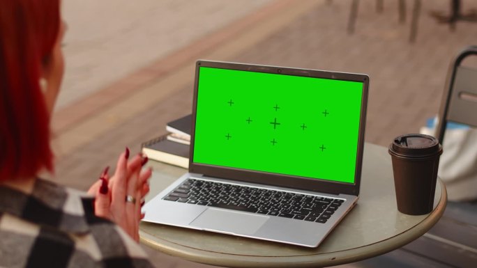 年轻迷人的红发女孩在学习期间用一台绿屏模拟笔记本电脑进行视频通话，当时她正坐在户外咖啡馆里喝咖啡，从