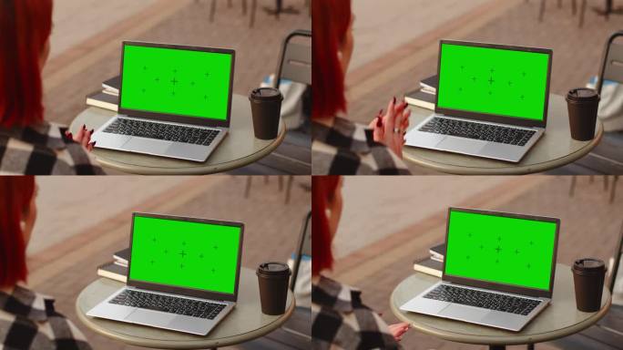年轻迷人的红发女孩在学习期间用一台绿屏模拟笔记本电脑进行视频通话，当时她正坐在户外咖啡馆里喝咖啡，从