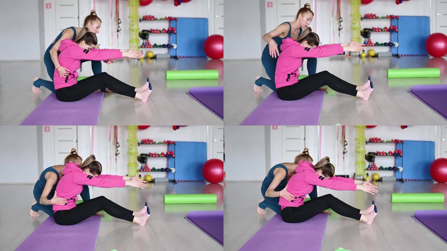 健身教练帮助一位年轻女子坐在健身房的瑜伽垫上做身体弯曲。摆脱背部疼痛的概念，恢复脊柱的健康。