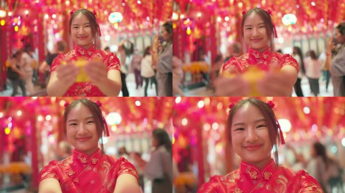 穿着红色旗袍的年轻亚洲女孩给你她手里拿着的金元宝。笑得很开心，看了看镜头。中国新年的概念。