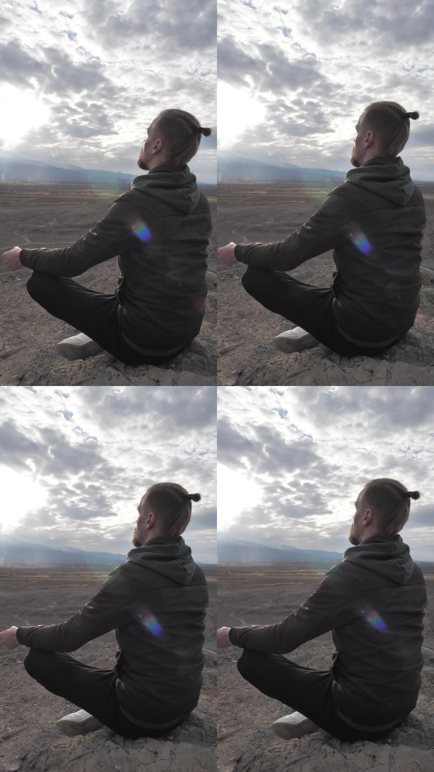 垂直:一个英俊的白种人扎着马尾辫，在日落时分坐在山上的一块石头上冥想。你内心的和谐与平衡。孤独的人在