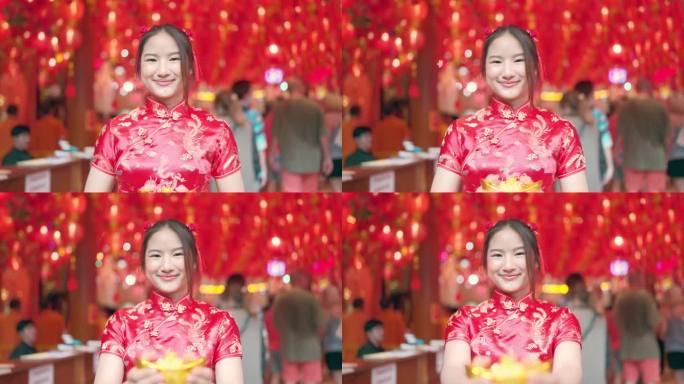 穿着红色旗袍的年轻亚洲女孩给你她手里拿着的金元宝。笑得很开心，看了看镜头。中国新年的概念。