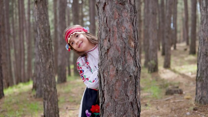 身着民族民间刺绣服装的年轻保加利亚女子躲在森林里的树干后面