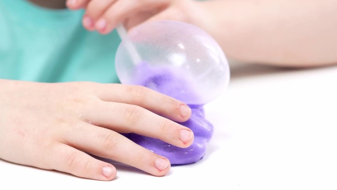 一个孩子玩着一个由亮紫色黏液制成的玩具，用管子吹泡泡，一个时髦的液体玩具粘在他的手上和手指上